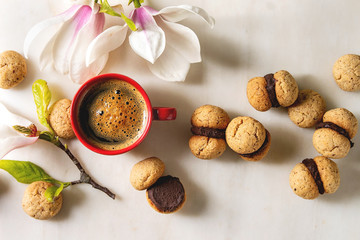 Fototapeta na wymiar Baci di dama homemade italian hazelnut biscuits cookies with chocolate cream served with red cup of espresso coffee and magnolia flowers over white marble background. Flat lay, space