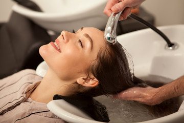 Happy beautiful woman smiling with her eyes closed while professional hairdresser washing her long healthy hair. Attractive woman enjoying relaxing hair treatment at beauty salon
