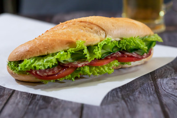 Fresh submarine sandwich with sausage, cheese, bacon, tomatoes, lettuce, cucumbers and onions on a dark wooden background. Mug of beer