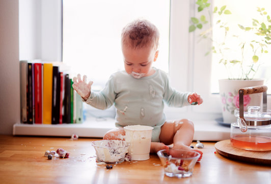 A Small Messy Toddler Boy Sitting On Kitchen Counter At Home, Eating.