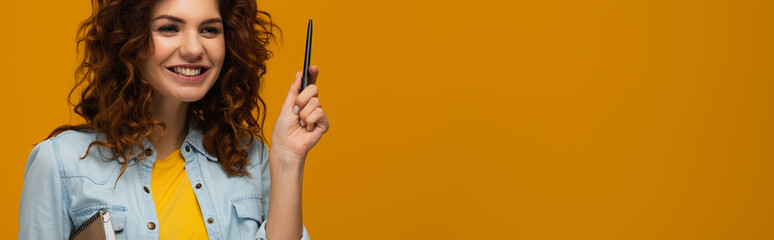 panoramic shot of smiling redhead young woman holding notebook and pen on orange