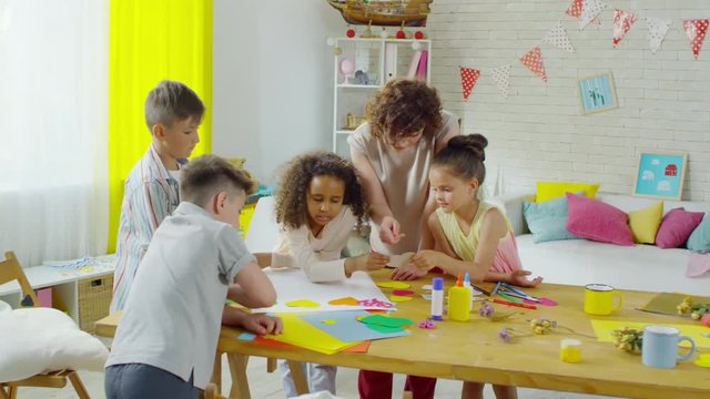 Multi-ethnic group of cute little kids applying colorful hearts to paper while making craft work together with female teacher in kindergarten