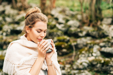 Beautiful woman holding cup of coffee or tea, covered with beige big scarf, posing outdoors