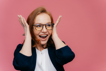 Positive smiling redhead woman covers ears, happy to hear something noisy and funny, wears transparent glasses, black and white elegant clothes, stands over pink background with free space for text
