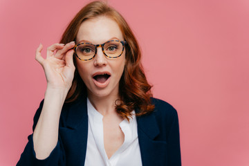Headshot of surprised red haired woman keeps hand on frame of glasses, jaw dropped, can not believe her eyes, wears elegant suit, shocked by unexpected rumor, stands over pink studio wall, free space