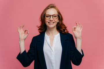 Pleased female financier makes okay gesture with both hands, has pleasant smile on face, wears...