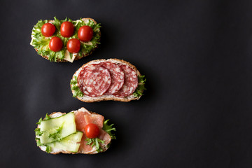 Fresh sandwiches with sausage, cheese, bacon, tomatoes, lettuce, cucumbers on a black background....