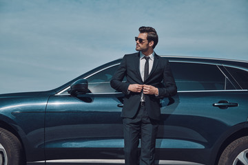 Always on style. Handsome young businessman adjusting jacket and smiling while standing near his car outdoors