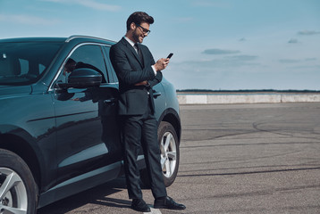 Plakat Confident manager. Full length of handsome young businessman using smart phone while standing near his car outdoors