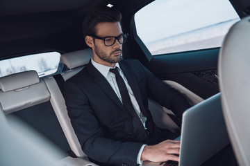 Young and successful. Handsome young man in full suit working using laptop while sitting in the car