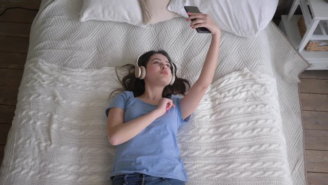 merry woman into headphones enjoy music and takes selfie photo on mobile phone while relaxing on bed, top view