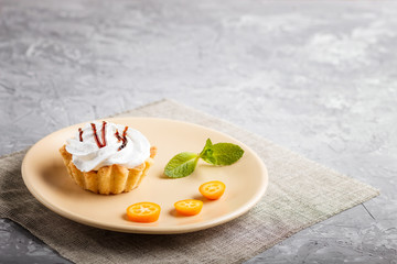 Cake with whipped egg cream on a light brown plate with kumquat slices and mint leaves on a gray concrete background. side view, copy space.