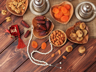 Ramadan concept. Delicious dates, dried figs, dried apricots, walnuts, almonds, raisins on bamboo and silver plates, rosary beads with Islamic holy book Quran on a pine wooden table background.