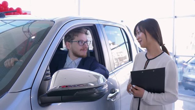 portrait of car sales manager with buyer young couple who shows keys inside machine while buying new family vehicle at auto salon