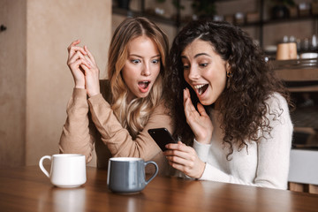 Emotional shocked girls friends sitting in cafe using mobile phone.