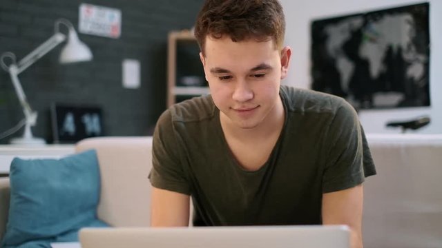 A teenage caucasian boy is happy working on the laptop in his room and nods his head
