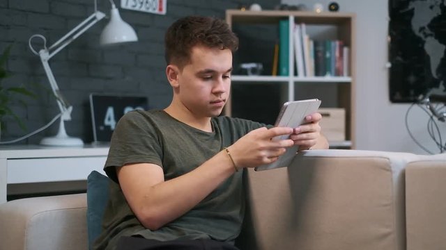 Portrait view of a young guy sitting on the couch and sufring the web on his tablet