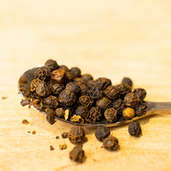 Whole Black Pepper Corns on Metal Spoon on Wooden Chopping Board.