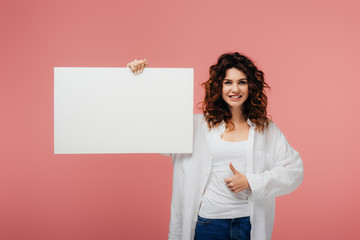 happy curly woman with red hair holding blank placard and showing thumb up on pink