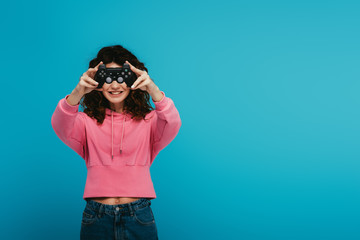 happy curly redhead girl covering face with joystick on blue