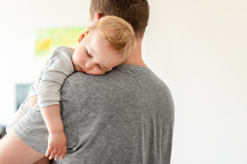 Portrait of cute adorable blond caucasian toddler boy sleeping on fathers shoulder indoors. Sweet...