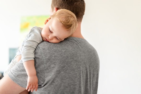 Portrait of cute adorable blond caucasian toddler boy sleeping on fathers shoulder indoors. Sweet little child feeling safety and care on daddys hand. Single father concept