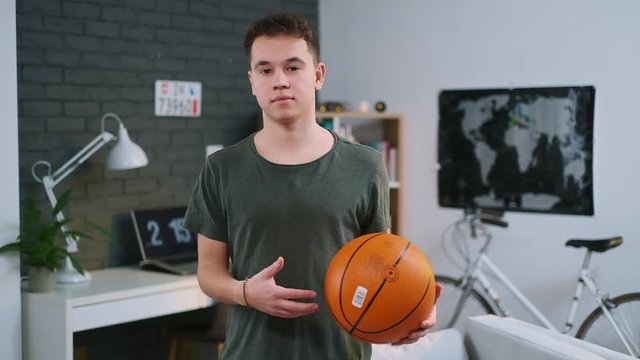 A handsome teenager passing the basketball ball from one hand to another in his room, front view