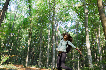 Naklejka na ściany i meble Backview of young woman jumping happy in the air, taking a walk in the forest, carrying a backpack in the forest on sunset light in the autumn season.