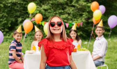 childhood, valentine's day and summer concept - smiling preteen girl with heart shaped sunglasses over birthday party at park