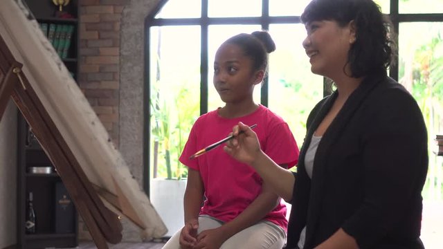 Young teacher giving African American kid an art lesson and teaching how to paint on easel inside living room in natural ambient light