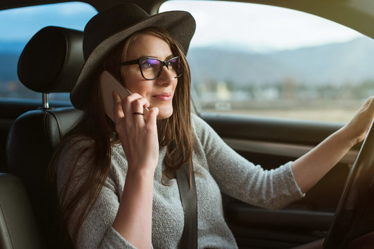 Young hipster woman with eyeglasses and hat driving the car and holding smartphone