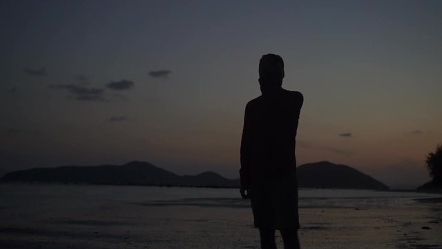 Young handsome man silhouette on the tropical beach sunset 4k.  Athletic male raises head and touches hair slow motion. Horizon with mountains and ocean on the back.
