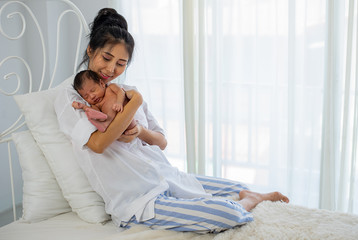 Obraz na płótnie Canvas Asian white shirt mother holds her little sleeping newborn baby on her chest and sit on white bed in front of glass windows with white curtain