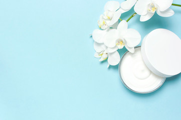 Beauty Spa concept. Opened plastic container with cream and White Phalaenopsis orchid flowers on blue background Flat lay top view. Herbal dermatology cosmetic hygienic cream, organic cosmetic Natural