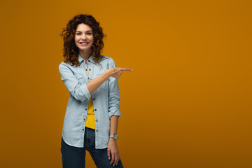 Obraz premium cheerful curly redhead woman standing and pointing with finger on orange