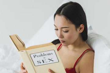 selective focus of concentrated asian woman reading problems in sex book while lying in bed