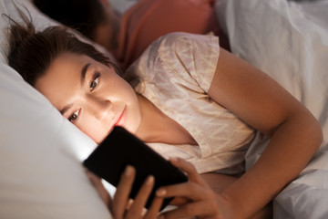Obraz na płótnie Canvas technology, internet addiction and cheat concept - woman using smartphone at night while her boyfriend is sleeping