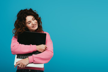 cheerful curly redhead girl hugging laptop with blank screen while standing on blue