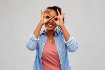 people, gesture and portrait concept - happy african american young woman looking through finger glasses over grey background