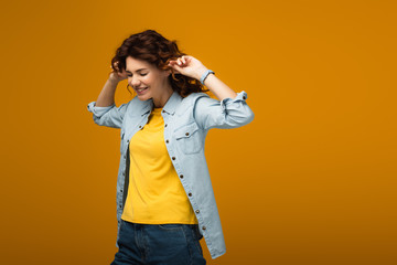 cheerful redhead woman plugging ears while standing on orange