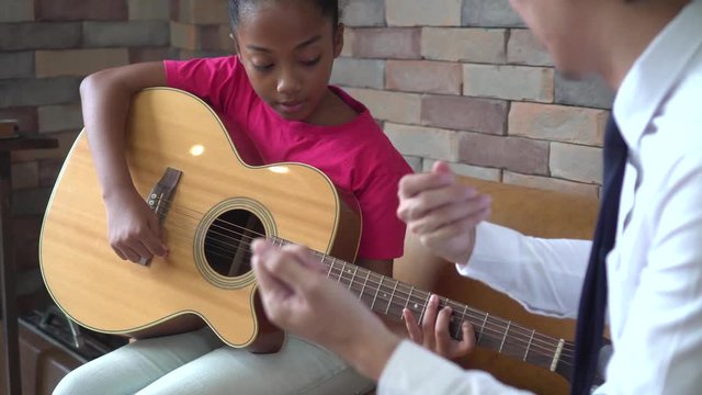 Young Asian male teacher giving a guitar lesson and teaching how to play guitar to African American girl in indoors room