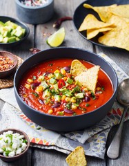 Mexican tomato, bean, bell pepper soup in black bowl with nachos chips. Wooden background.