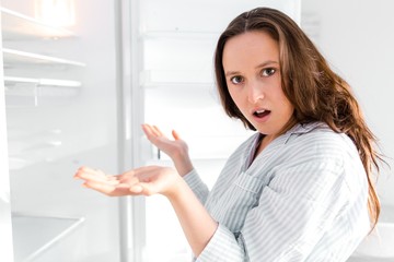 Surprised girl standing in the kitchen looks into the empty refrigerator spreading his hands to the...