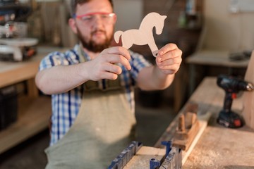 Bearded carpenter holding a wooden toy made a horse in the home workshop