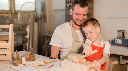 Father and son in the carpenter's shop make a wooden toy tank. Paternal care. Teaching a child craft
