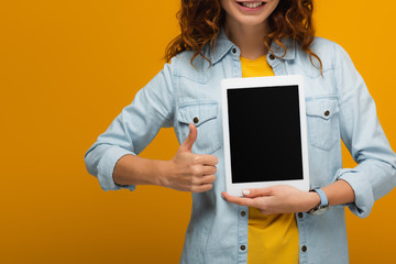 cropped view of cheerful curly woman holding digital tablet with blank screen and showing thumb up isolated on orange