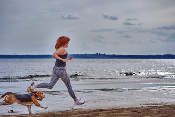 A red-haired middle-aged woman in sportswear runs along the sandy shore of a large river with her...