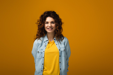 happy curly redhead woman looking at camera and smiling on orange