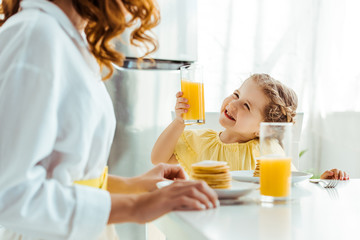 selective focus of emotional happy daughter looking at mother and holding glass with orange juice...