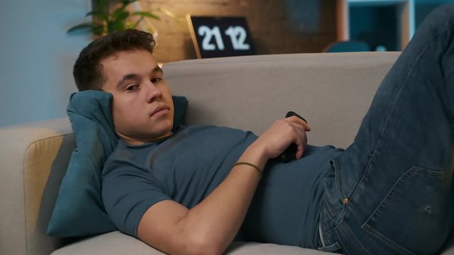 A teenage boy is lying on the comfortable couch and watching TV, he's bored, so he switches channels with the remote control.